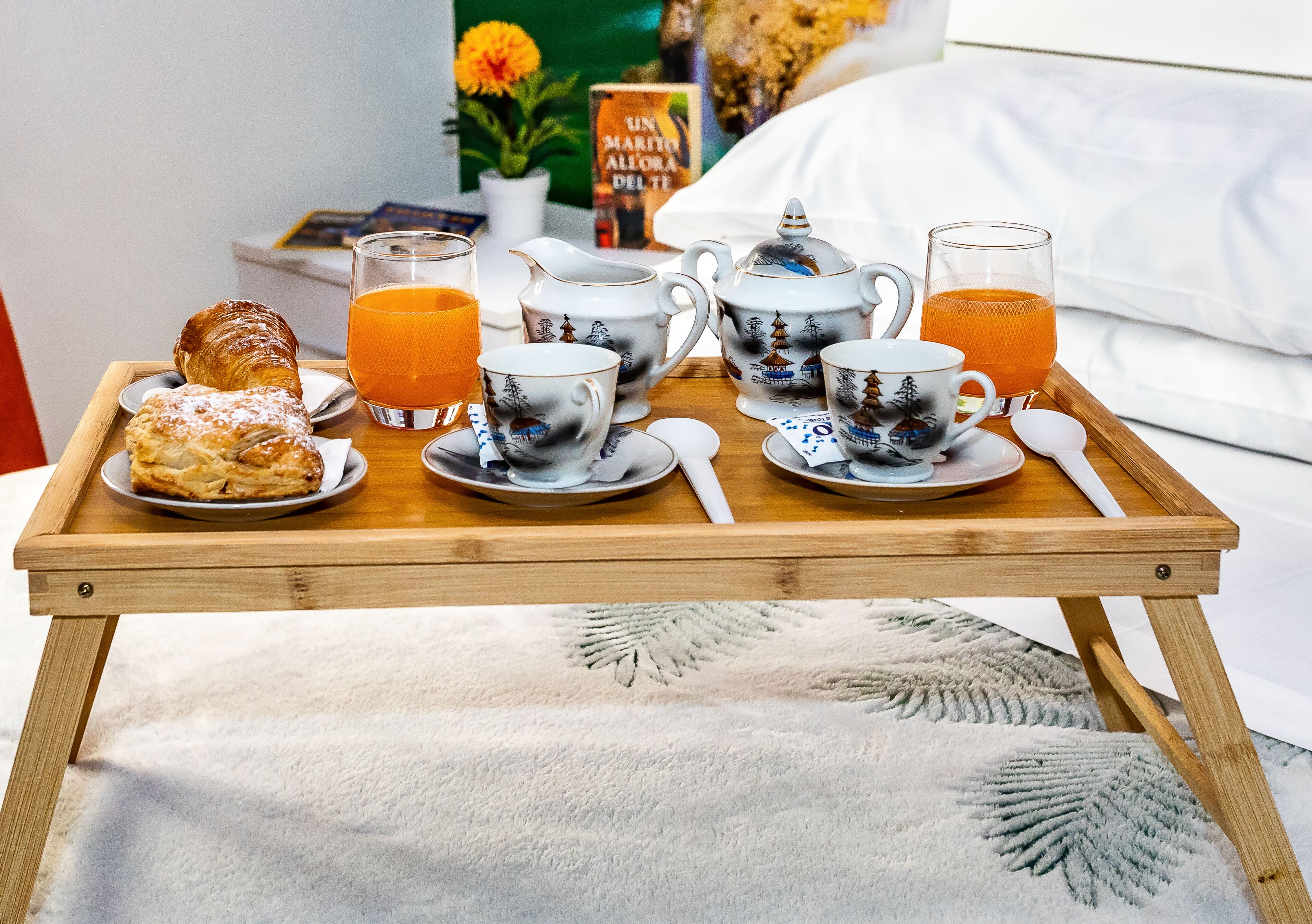 Bandeja de desayuno con croissant, tetera, tazas y zumo de naranja sobre una cama.