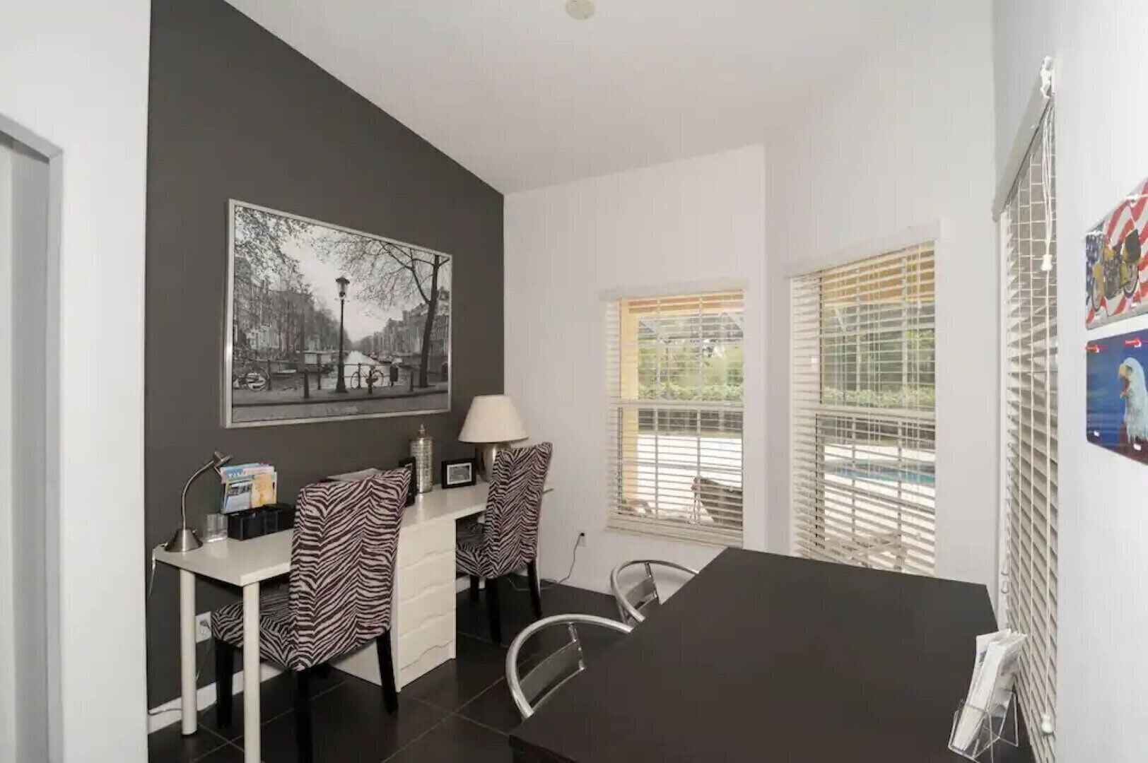A modern room with a black desk, two zebra-patterned chairs, a black sofa, a lamp, and a large black-and-white cityscape painting on the wall.