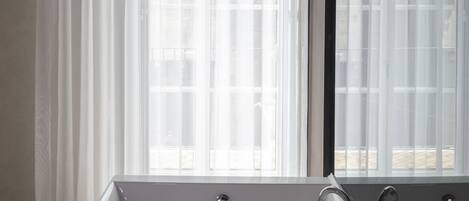 A modern bathroom with a freestanding bathtub, a large mirror, and a window with sheer curtains.