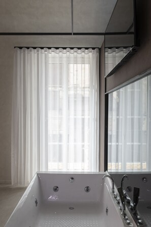 A modern bathroom with a freestanding bathtub, a large mirror, and a window with sheer curtains.