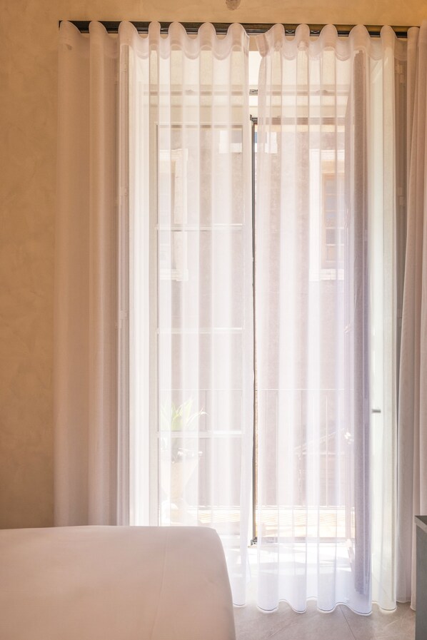 A room with a bed, a window with sheer curtains, and a view of the outside.