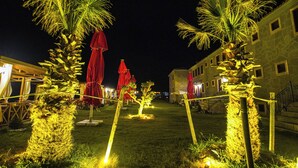 A nighttime outdoor area with palm trees, red umbrellas, and a building in the background. - Bakbay Bozcaada (Bozcaada)