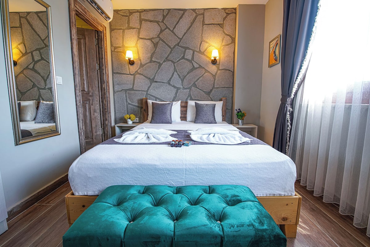 A bedroom with a stone wall, a bed with a white and teal bedspread, two bedside lamps, a wooden bench, and a mirror.