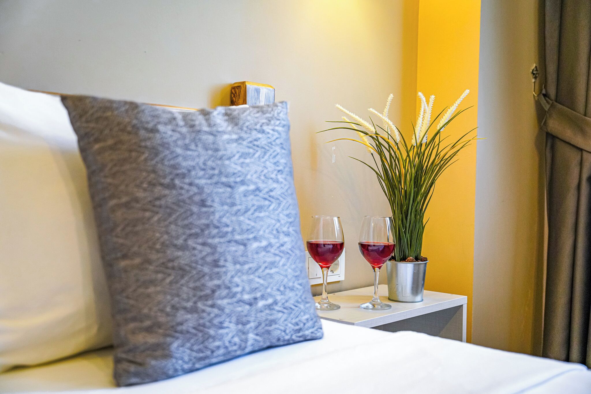 Una cama bien tendida con un cojín estampado, una mesita de noche con dos copas de vino y una planta en maceta, y una puerta con cortinas.