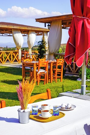 An outdoor dining area with tables and chairs, a white tablecloth, and a view of the surrounding landscape. - Bakbay Bozcaada (Bozcaada)