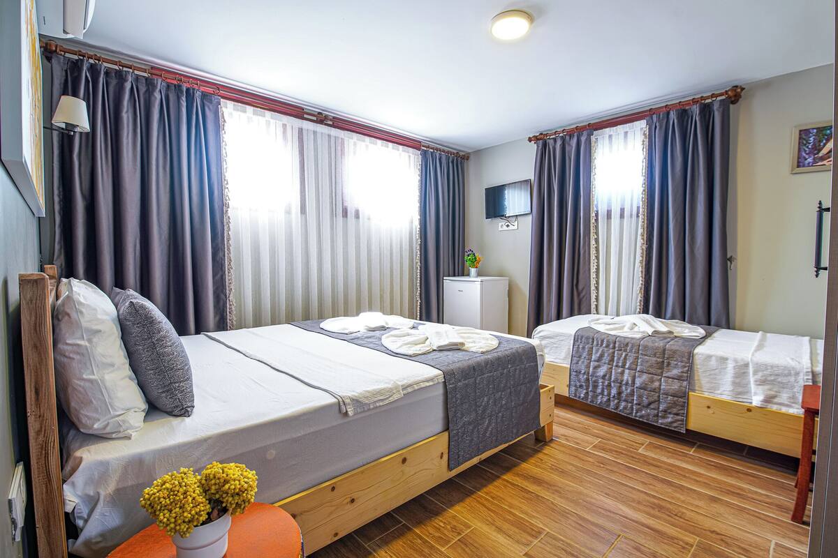A room with two single beds, a small table with a vase of flowers, a television on the wall, and a window with curtains.