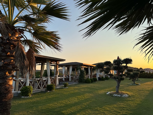 A covered outdoor seating area with tables and chairs, surrounded by palm trees and a well-maintained lawn. - Bakbay Bozcaada (Bozcaada)