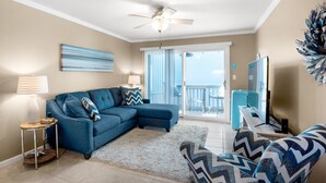A living room with a sectional sofa, a small side table, a floor lamp, a ceiling fan, a large window, and a balcony with a view. - 🌴Steps2Beach-Community Pool-2BR Villas on the Gulf A8🌴 (Pensacola Beach)