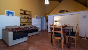 A cozy living room with a sofa, a dining table with chairs, a ceiling light, and shelves with decorative items. - Casa Nora T2 (Castro Marim)