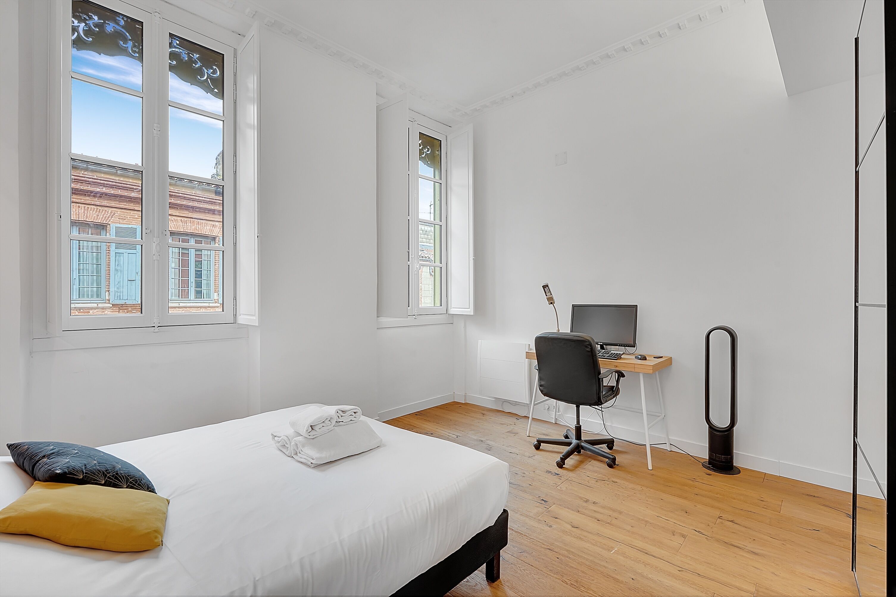 A bedroom with a bed, a desk with a computer, a chair, and a standing heater.