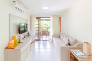 A modern living room with a sofa, a coffee table, and a TV mounted on the wall. There is a balcony with a view of greenery. - PF35 Ótimo Quarto e Sala a 25m da Praia (Mata de São João)