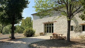 Ein Steinhaus mit Kiesweg, einem Holztisch und Bänken, und ein Baum im Vordergrund.