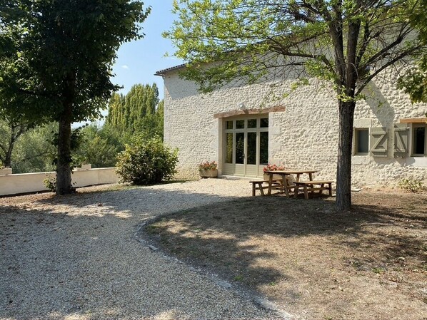 A stone building with a gravel driveway, a wooden table and benches, and a tree in the foreground.