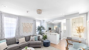 A modern living room with a grey sectional sofa, a round coffee table, and a blue armchair. There is a kitchen area with light blue cabinets and a white countertop. - Hot Tub Oasis • Parking • 1 Block to King St! (Charleston)