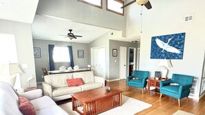 Ein großzügiges Wohnzimmer mit hoher Decke, Holzboden, Deckenventilator, einem weißen Sofa, einem Holztisch und Teakfarbenen Sessel. Ein Gemälde hängt an der Wand.
