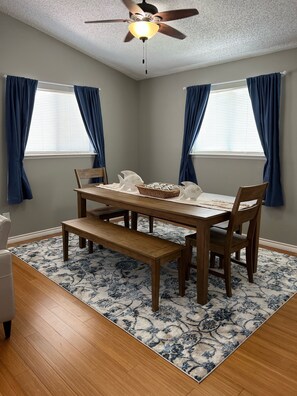 Comedor con mesa de madera, sillas, ventilador de techo y alfombra estampada.