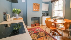 Una cocina moderna con una placa de inducción negra, armarios blancos y un mueble de madera sobre la estufa. Hay una mesa de comedor con sillas naranjas y un tapete estampado.
