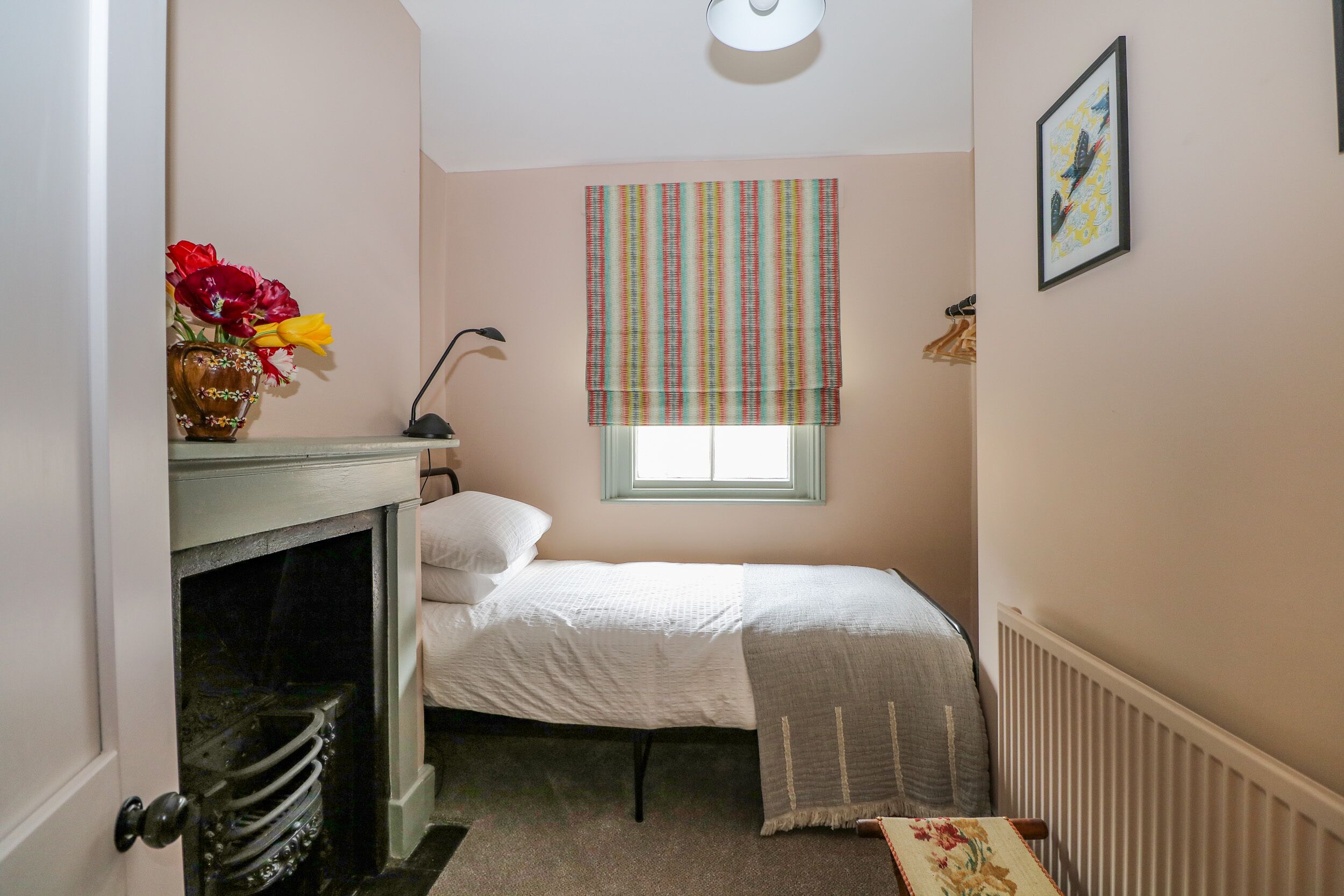 Ein kleines Schlafzimmer mit einem Bett, einem Heizkörper, einem Fenster mit gestreiften Vorhängen, einem gerahmten Bild an der Wand und einer Vase mit Blumen auf einem Tisch.