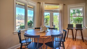 A dining area with a round wooden table, six chairs, and large windows offering a view of the outdoors.