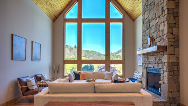 A spacious living room with a large window, a stone fireplace, and a wooden ceiling.