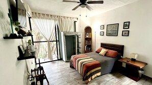 A bedroom with a bed, a ceiling fan, a bookshelf, a window with curtains, and a small table with a lamp.