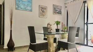 A dining area with a glass-top table, four wicker chairs, a vase, and two framed artworks on the wall.
