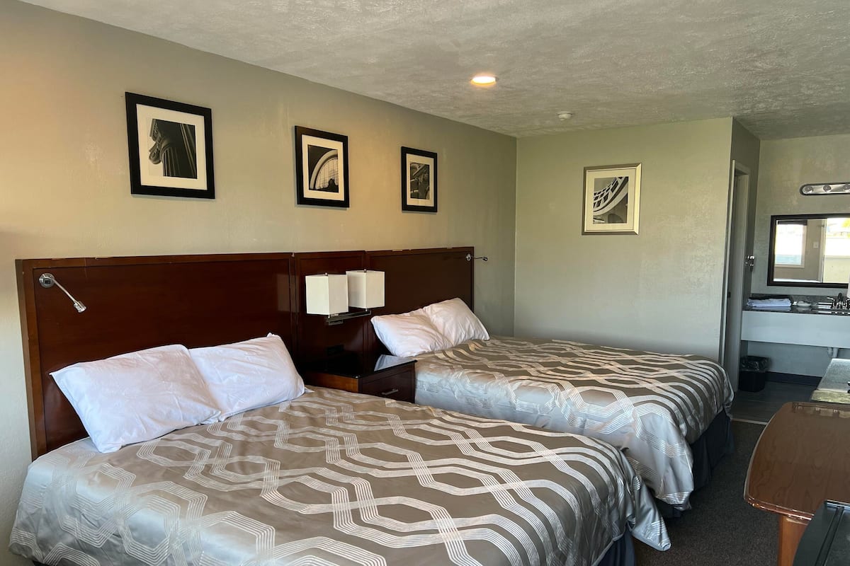 Une chambre d’hôtel avec deux lits, une tête de lit en bois, une table de chevet, un bureau, un miroir et des tableaux encadrés accrochés au mur.