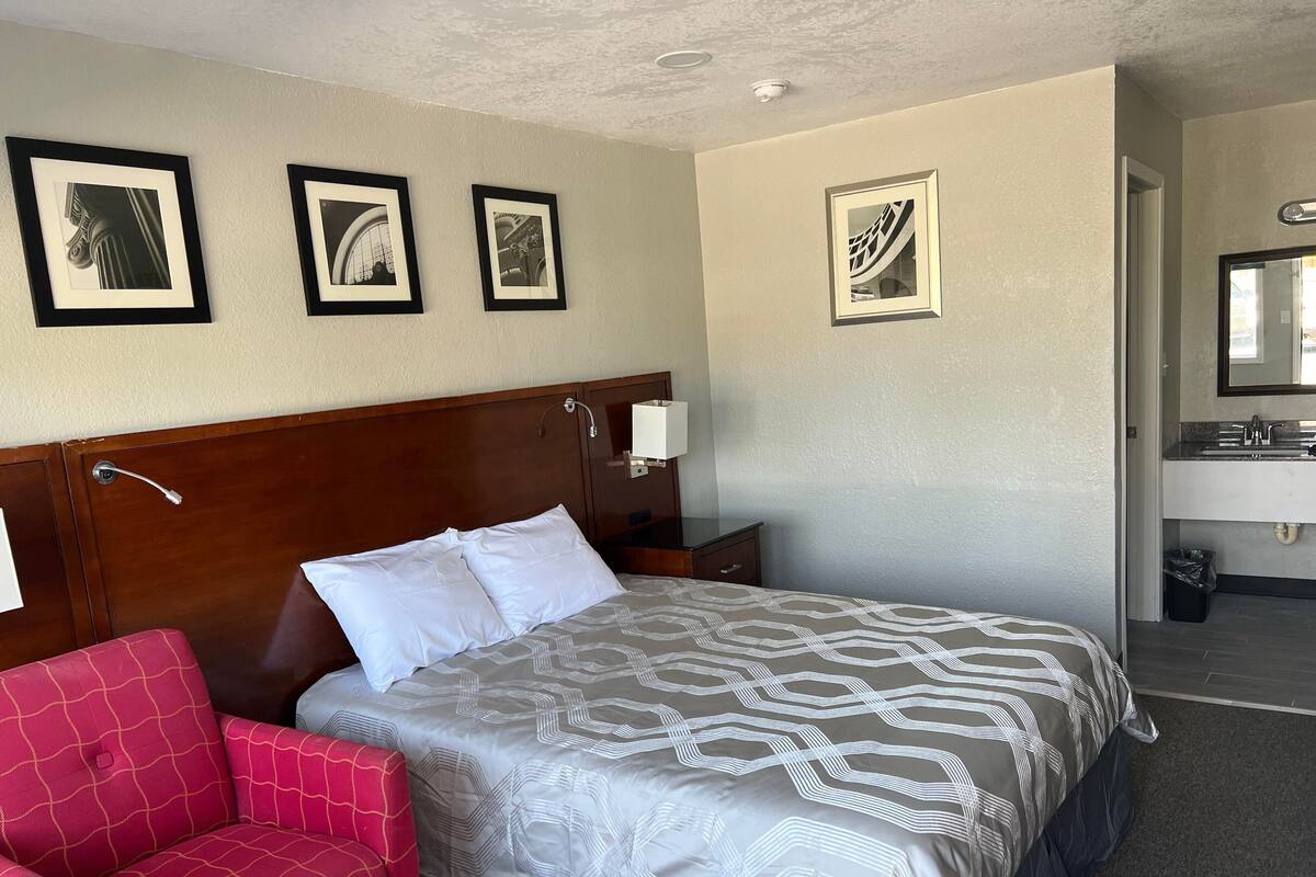 Une chambre d’hôtel avec un lit, une table de chevet, une lampe, une chaise et des tableaux encadrés au mur.