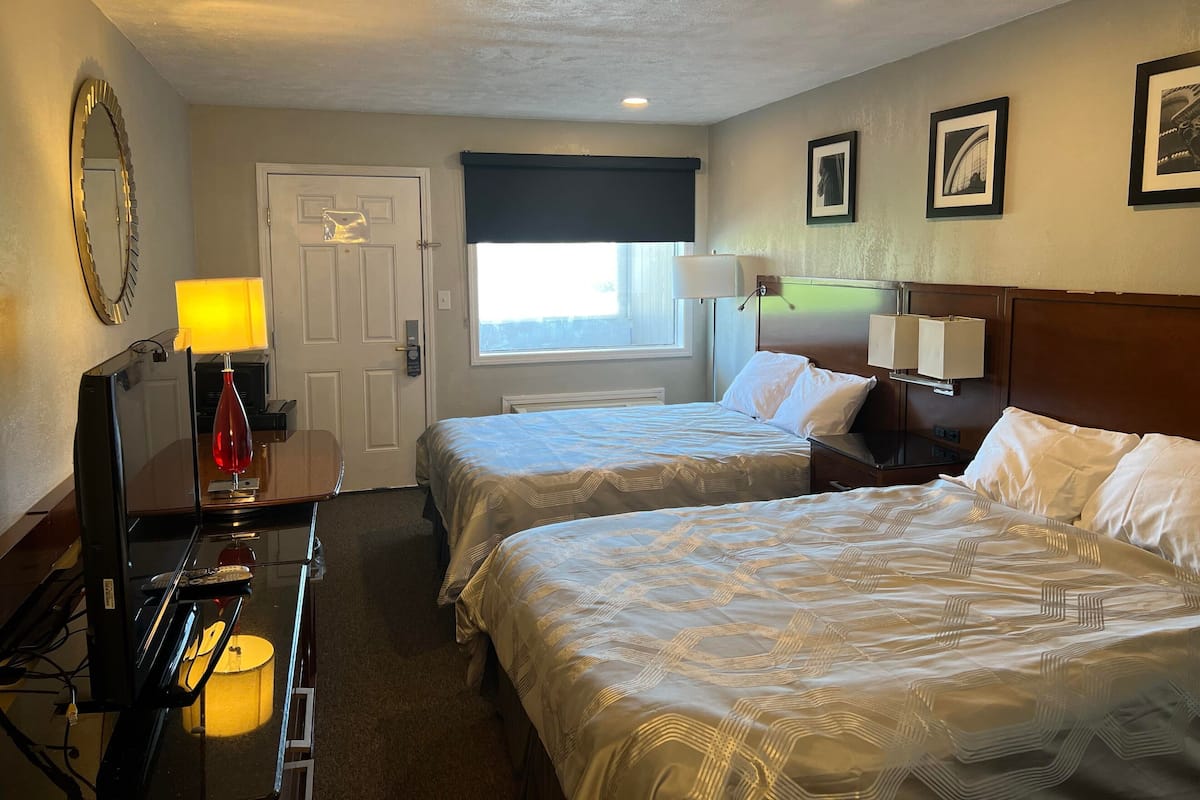 Une chambre d’hôtel avec deux lits, un bureau avec une lampe, un miroir, une fenêtre donnant sur un paysage agréable et des tableaux encadrés accrochés au mur.