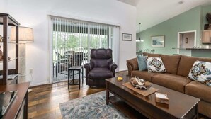 A living room with a brown leather sofa, a dark leather armchair, a wooden coffee table, and a patterned rug. There is a dining area with a table and chairs visible through a window with vertical blinds.