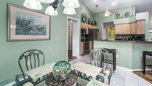A dining area with a glass table, chairs, and a chandelier. There is a kitchen in the background with cabinets and appliances.