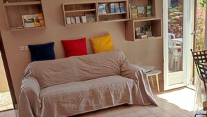 A room with a beige sofa, a small round table, and built-in wooden shelves with books and decorative items. - Quiet gite with garden 300 m from Le Guichet rer station (Orsay)