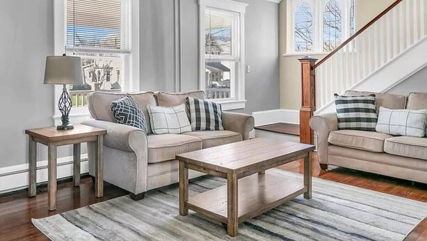 Una sala de estar con dos sofás, una mesa de centro y una lámpara. Hay ventanas con persianas y una escalera al fondo.