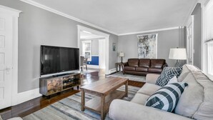 Una sala de estar con una televisión de pantalla plana, un sofá de cuero marrón, una mesa de centro de madera y un sofá de color claro con almohadones estampados.