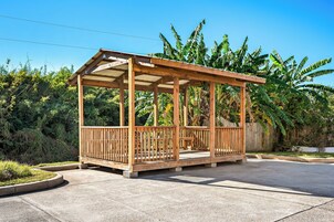 A wooden gazebo with a roof and railings, surrounded by a concrete patio and lush greenery.