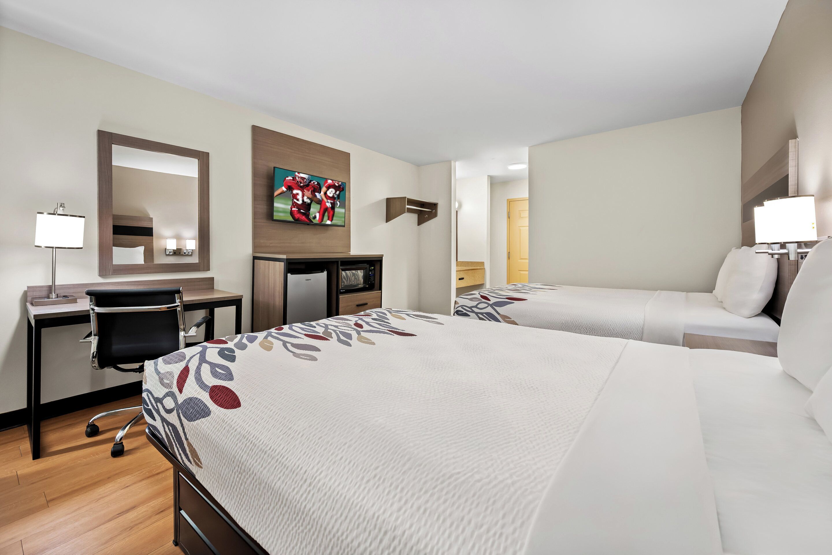 Habitación de hotel con dos camas, un escritorio con silla, televisor y espejo.