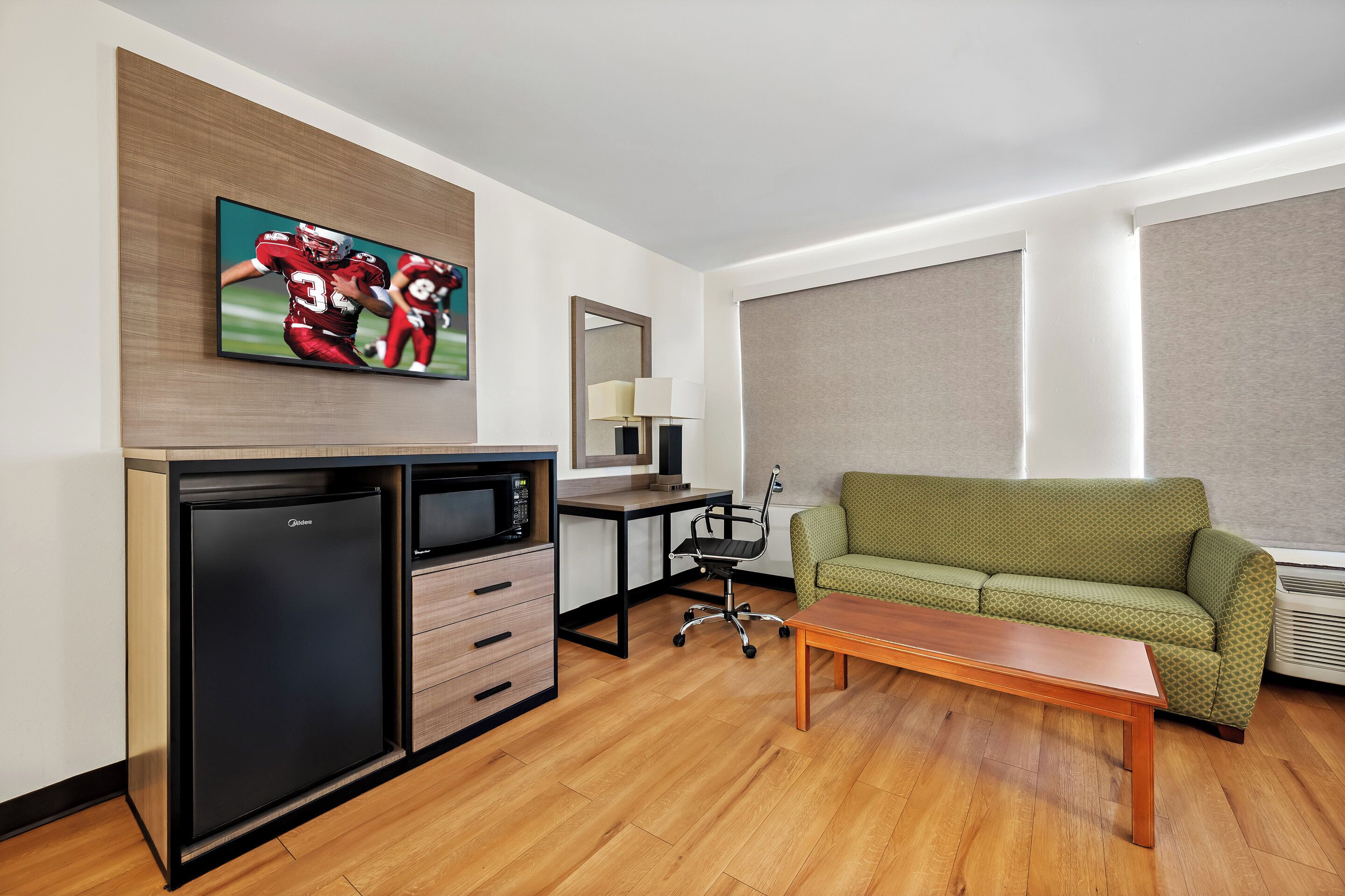 A compact hotel room with a kitchenette, a green sofa, a wooden coffee table, a flat-screen TV, and a large window with blinds.