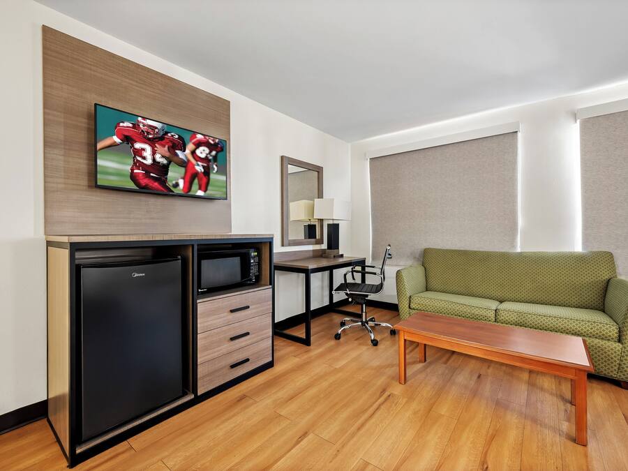 A compact hotel room with a kitchenette, a green sofa, a wooden coffee table, a flat-screen TV, and a large window with blinds.