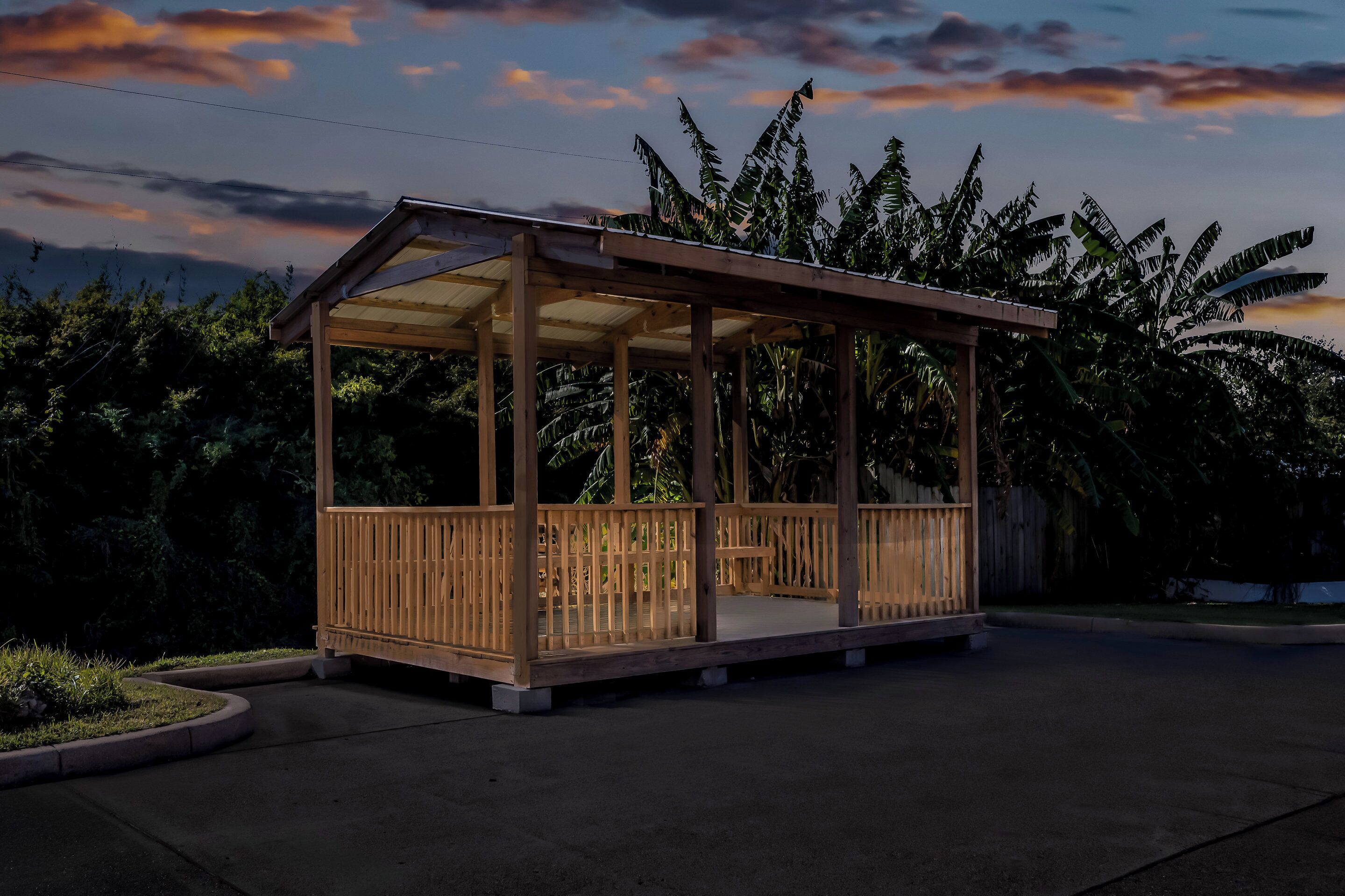 Ein hölzerner Pavillon mit Dach und offenen Seiten, umgeben von üppigem Grün und wolkenlosem Himmel.