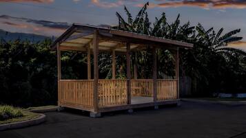 Ein hölzerner Pavillon mit Dach und offenen Seiten, umgeben von üppigem Grün und wolkenlosem Himmel.