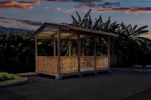 A wooden gazebo with a roof and open sides, surrounded by lush greenery and a clear sky.