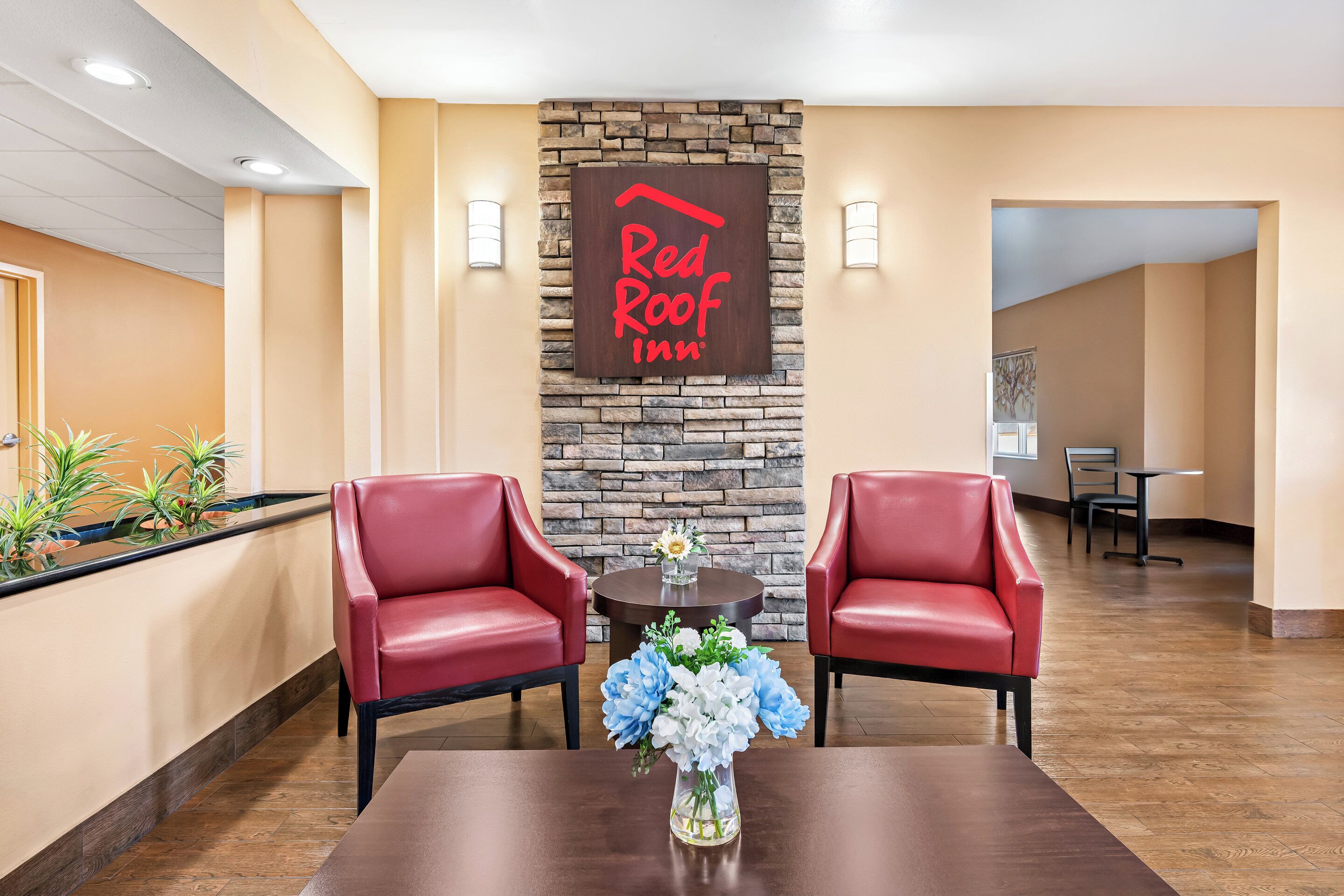 A Red Roof Inn reception area with two red leather chairs, a wooden table with a vase of flowers, and a stone wall with the inn's logo.
