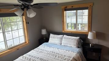 Un dormitorio con una cama, dos mesitas de noche, un ventilador de techo y una ventana con vistas a árboles.