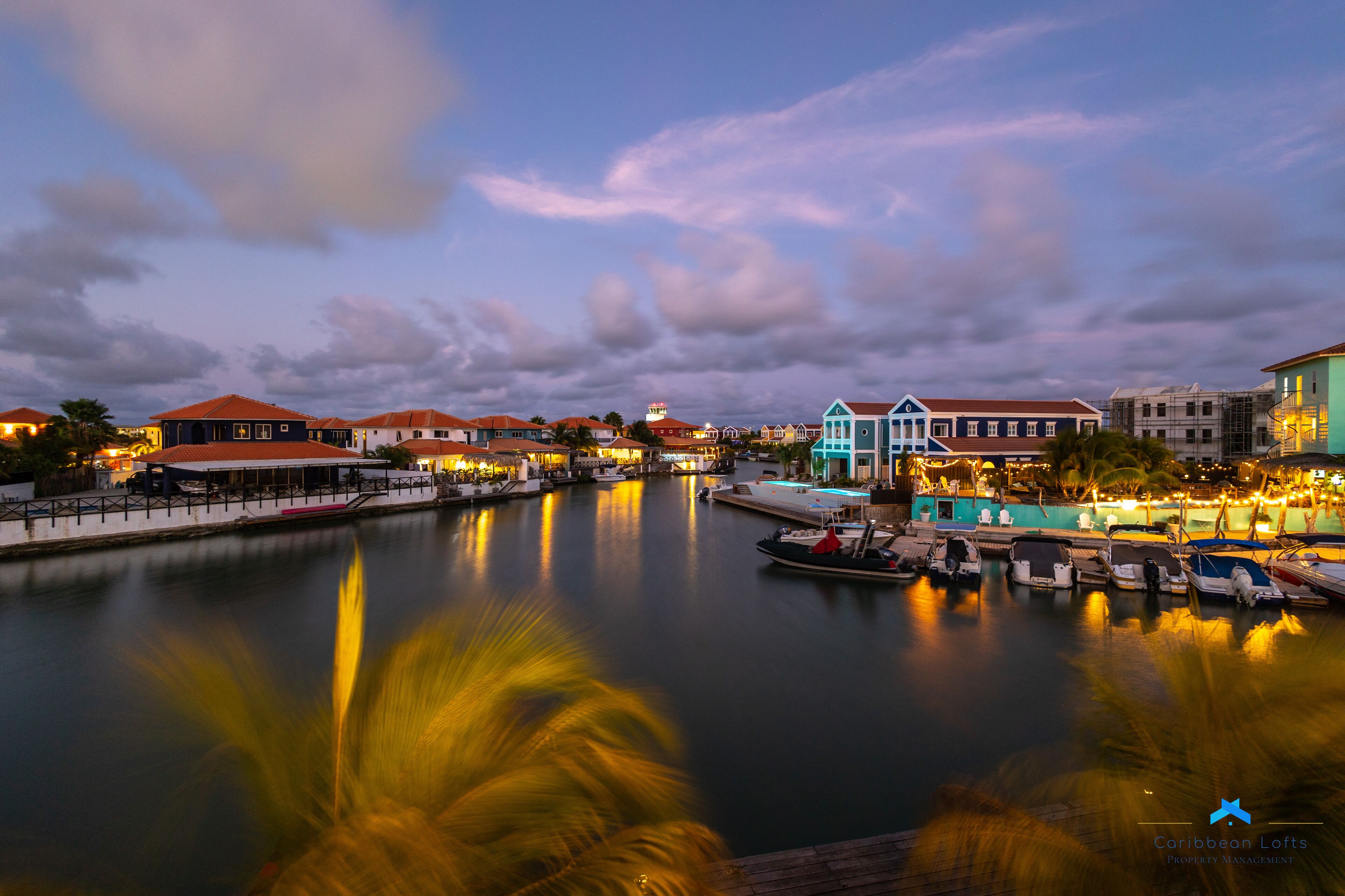 Uma marina com barcos atracados, edifícios à beira-mar e um corpo d'água calma que reflete as construções.