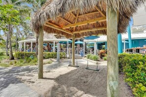 A covered outdoor area with a swing set, palm trees, and a building with blue accents in the background.