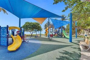 A playground with slides and climbing structures, shaded by blue canopies, and surrounded by trees.