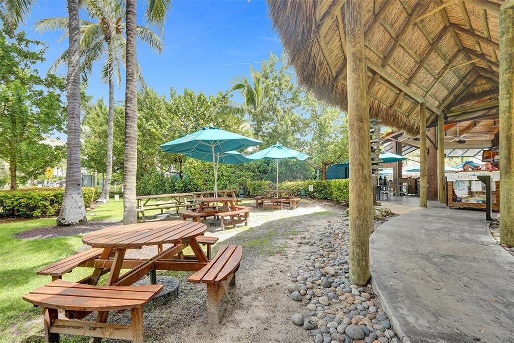 An outdoor patio area with wooden picnic tables and green umbrellas, surrounded by palm trees and a thatched-roof structure.