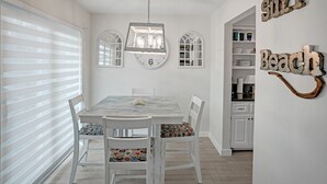 A dining area with a white table, chairs, and a chandelier. There are two arched windows and a wall-mounted clock.