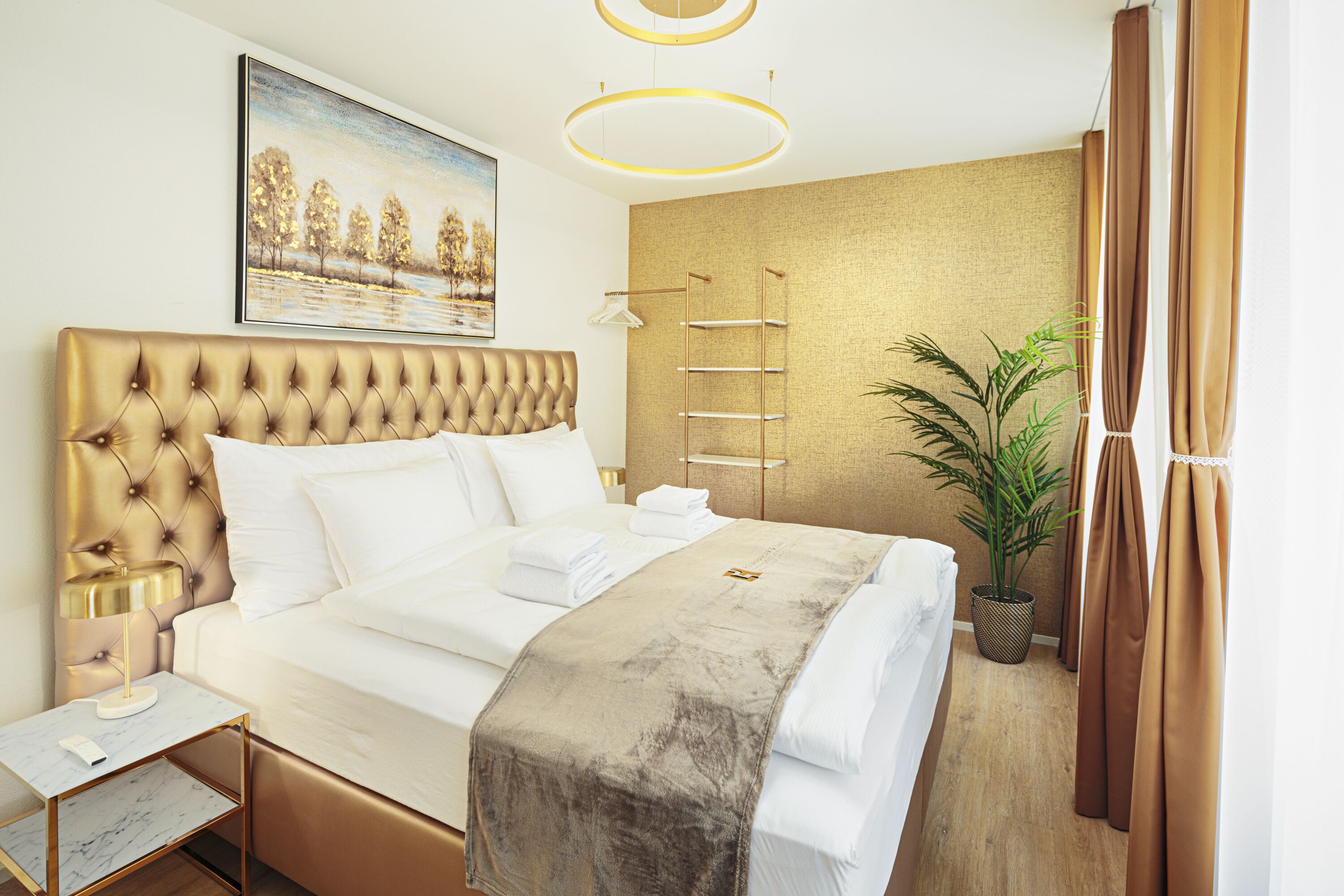 A hotel room with a tufted headboard, a large bed with white linens, a bedside table with a lamp, a painting on the wall, a plant, and a gold-colored wall.