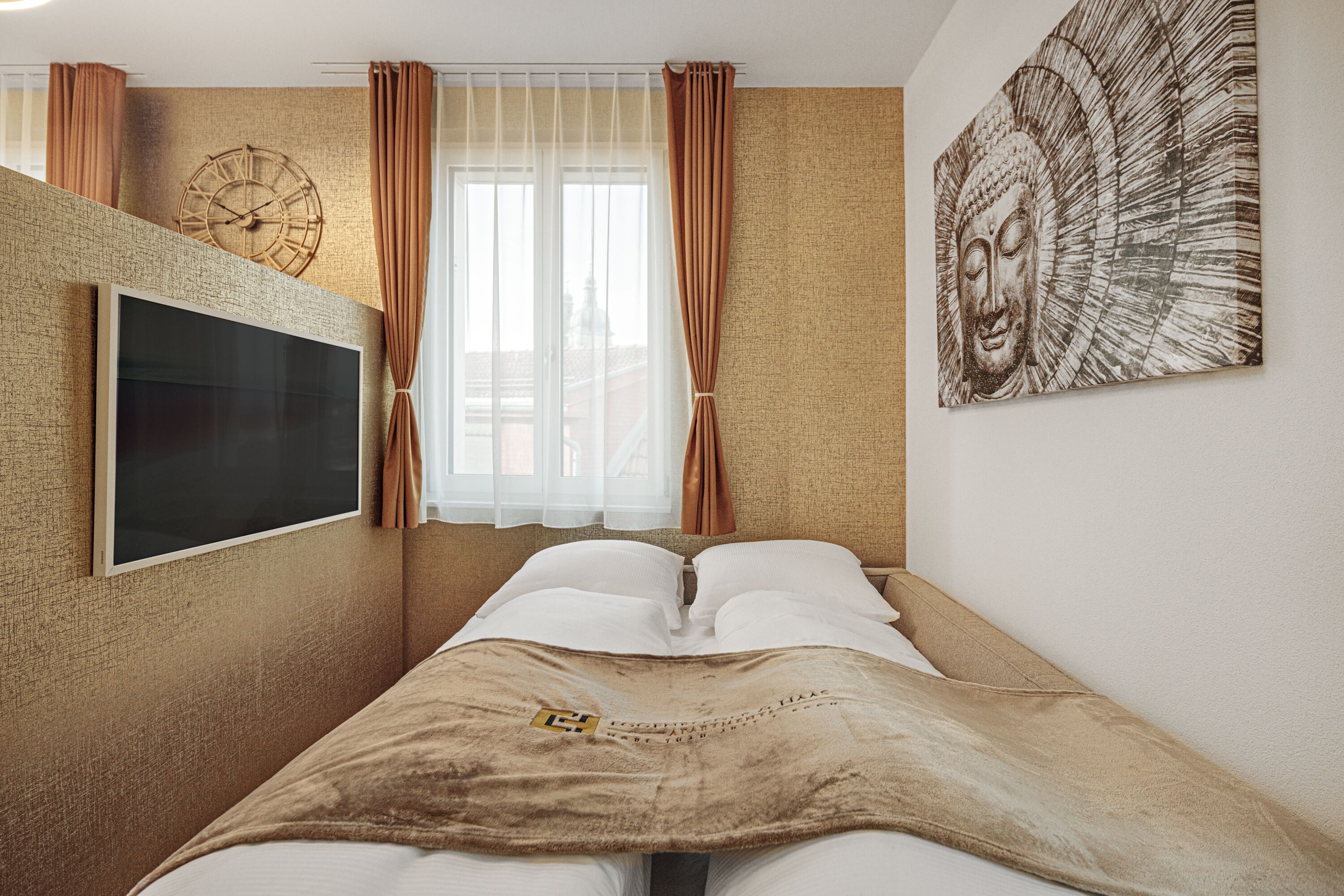 Une chambre à coucher avec un lit, un téléviseur fixé au mur, une horloge murale et un tableau représentant un Bouddha.